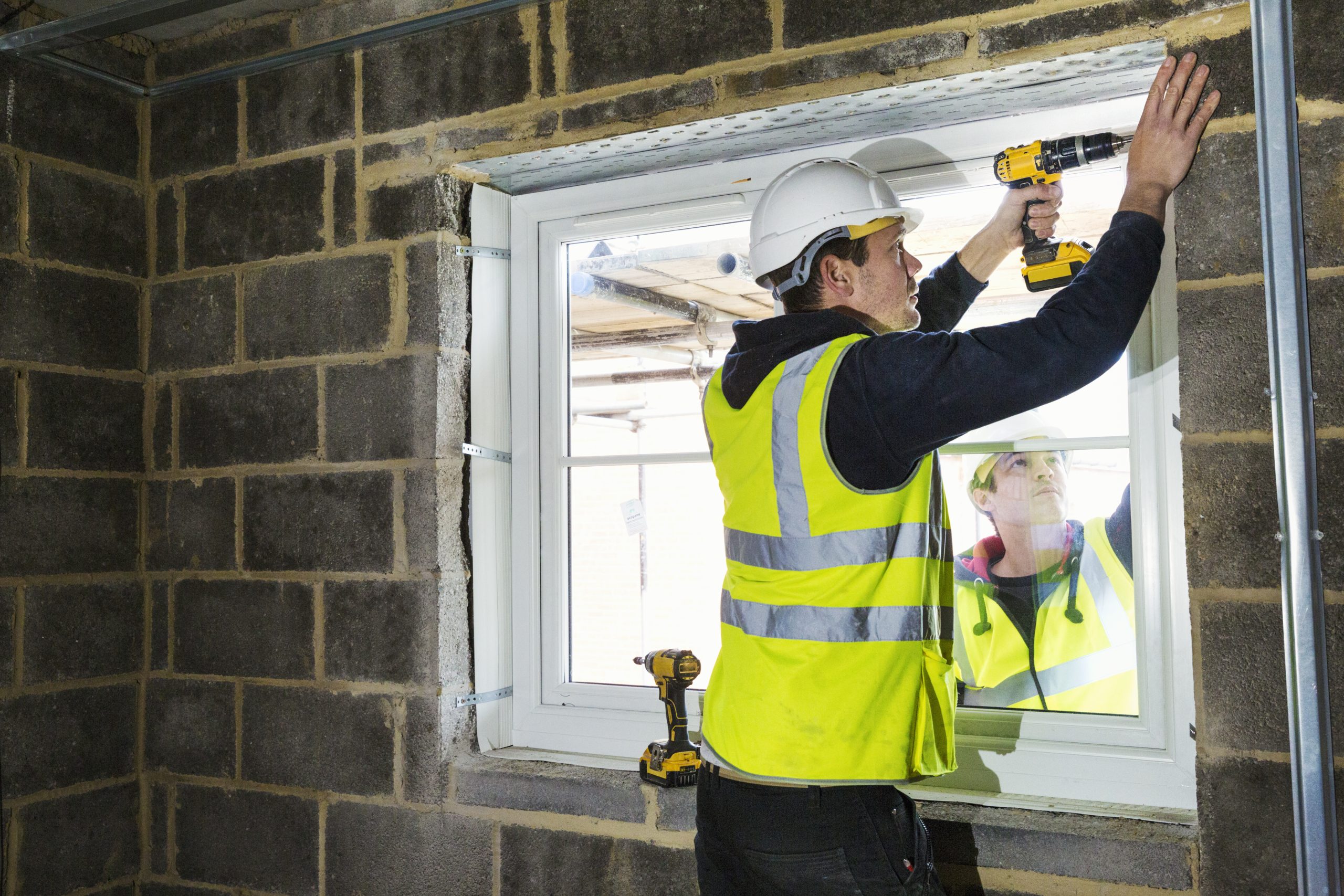 A workman on a construction site, a builder in hard hat using an electric drill on a window sill.,Construction Site,England
